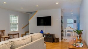 Ein Wohnzimmer mit einer Couch, einem an der Wand montierten Fernseher, einem Esstisch mit Stühlen und einer Treppe, die in einen anderen Raum führt.