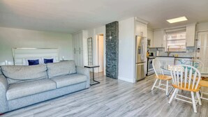 Una sala de estar moderna con un sofá gris, una mesa de comedor con sillas y una zona de cocina con armarios blancos y una pared de acento de piedra.