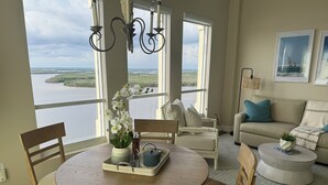 A dining area with a round wooden table, chairs, and a vase with white flowers. There is a view of a body of water through large windows. - Lovers Key Resort, Luxury Top Floor Penthouse with Best Views in the Resort (Fort Myers Beach)