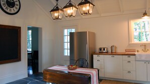 Eine Küche mit Kühlschrank, Herd und Spüle. Ein Holztisch ist mit einer Fruchtschale und einer rot-weißen Tischdecke gedeckt. An der Wand hängt eine Uhr, und über dem Tisch befinden sich drei Laternen.