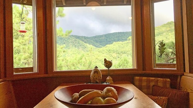 Una mesa de comedor de madera con un bol de pan y una figurita de pájaro. Se ven grandes ventanales con vistas a colinas verdes.