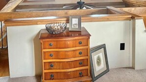 Un dormitorio tipo loft con techo inclinado, vigas de madera, tragaluces, ventilador de techo, cómoda de madera y un cuadro enmarcado.