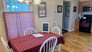 Un espace repas avec une nappe à carreaux, des chaises blanches, un panneau noir et une vue sur la cuisine.