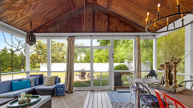 A covered patio with a wooden ceiling, large windows, a dining table with chairs, and a seating area with cushions.