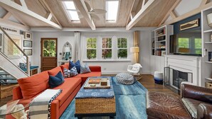 A spacious living room with a high ceiling, wooden beams, a fireplace, and a large window overlooking greenery.