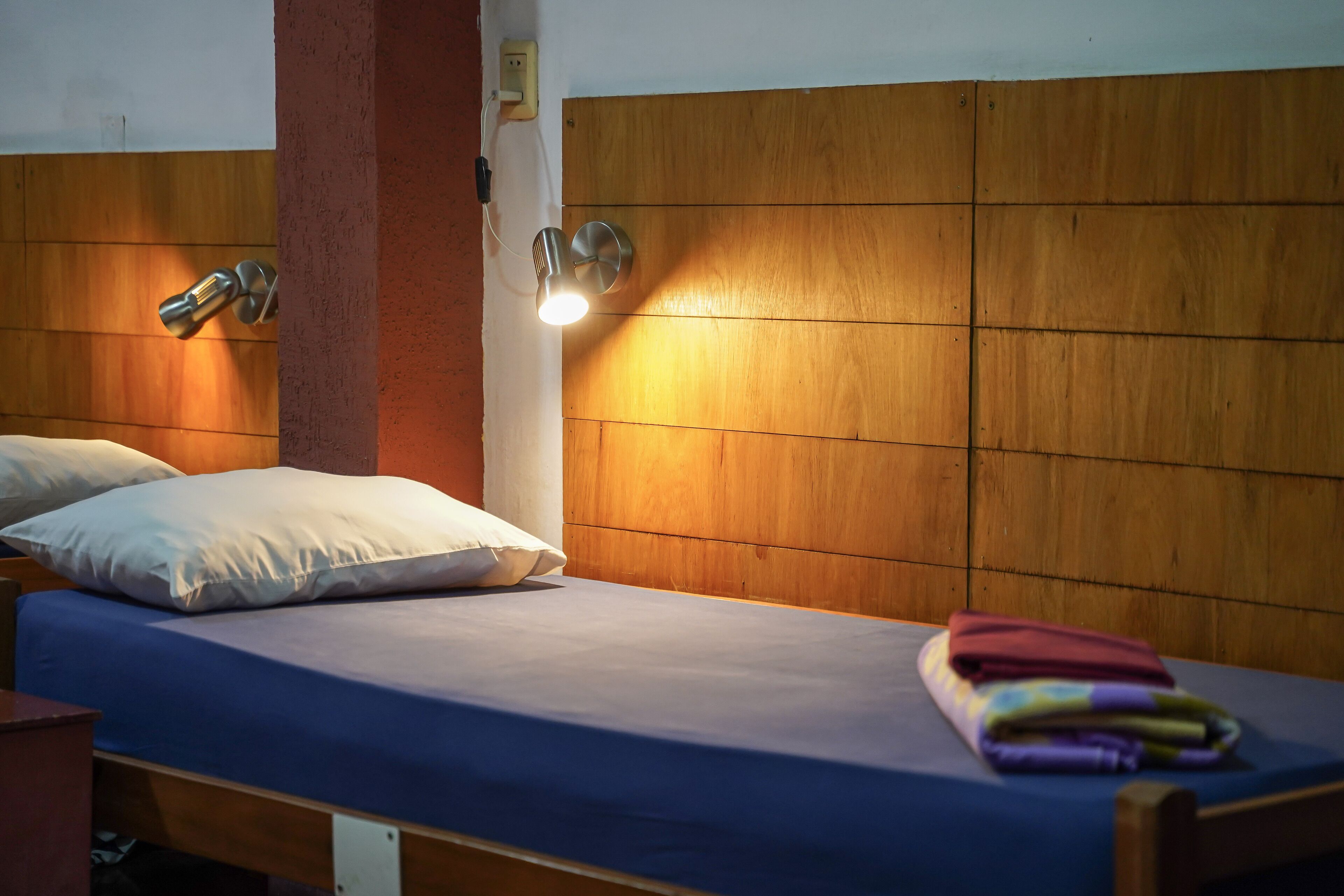 Ein Einzelbett mit blauer Matratze, zwei Kissen und einer Wandleuchte.