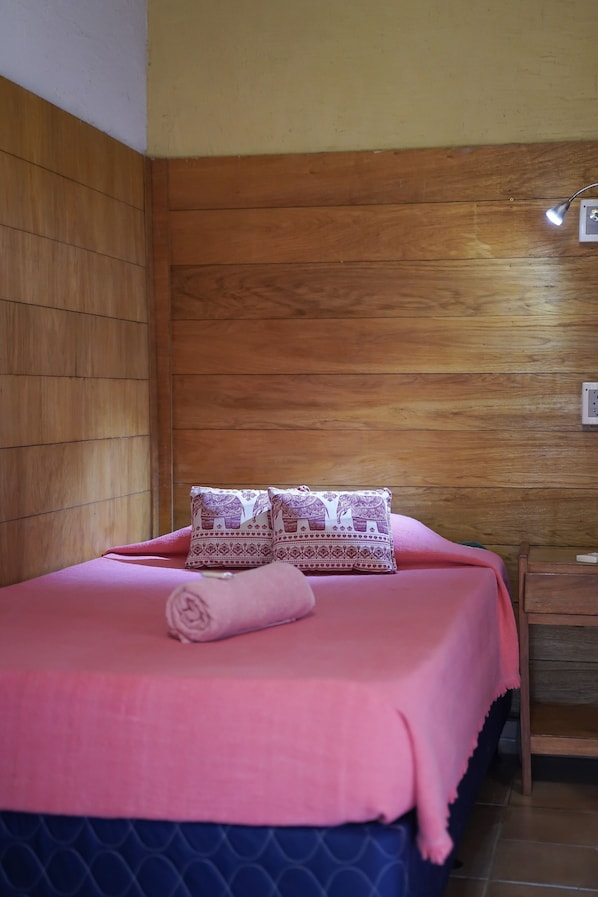Una cama con una colcha rosa y almohadillas estampadas, un cabecero de madera y una mesita de noche con una lámpara.