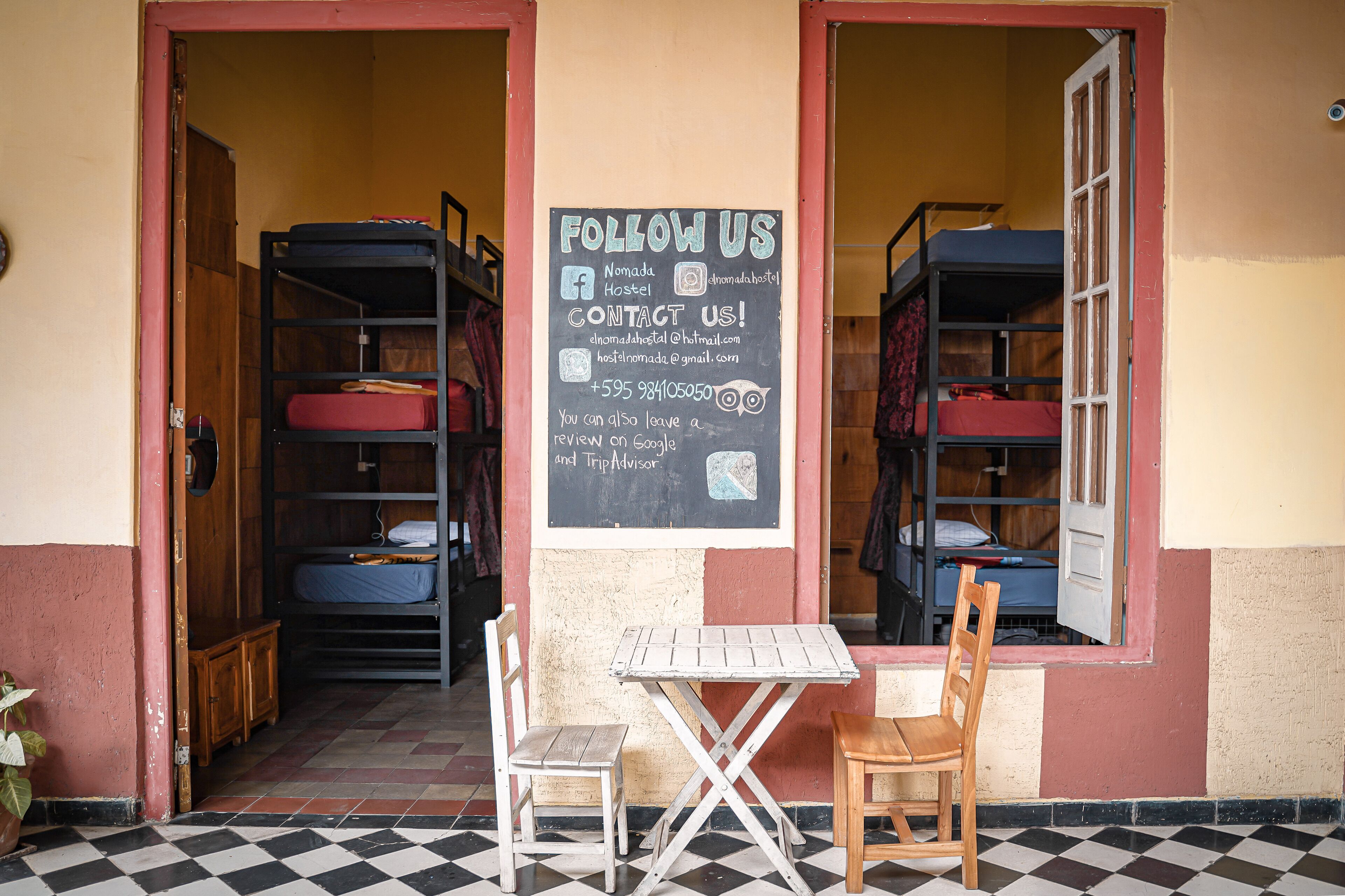 A hostel with bunk beds, a chalkboard sign advertising social media and contact information, and a small table with chairs outside.