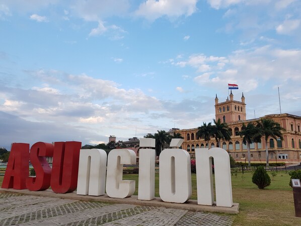 A large building with a prominent clock tower and flags, surrounded by a well-maintained lawn and palm trees. - El Nomada Hotel & Hostel (Asunción)
