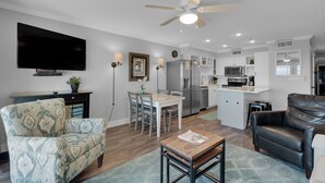 A modern living room with a sofa, a coffee table, and a TV. There is a dining area with chairs and a table. The kitchen has white cabinets and appliances. - BeachFront, Bch Svc, pool, wifi, Kitchen, grill (Destin)