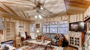 A rustic cabin interior with a wooden ceiling, a ceiling fan, a television, a fireplace, and a large window.