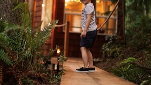 Niño de pie en una cubierta de madera frente a una cabaña rústica con luces colgantes.