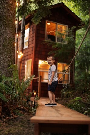 Un enfant se tient sur une terrasse en bois devant une cabane rustique, illuminée de guirlandes lumineuses.