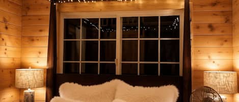 A cozy wooden cabin bedroom with a large bed, two bedside lamps, a ceiling fan, and string lights.