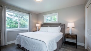 Une chambre avec un grand lit, deux tables de chevet avec des lampes, une fenêtre avec des rideaux et une vue sur les arbres à l’extérieur.