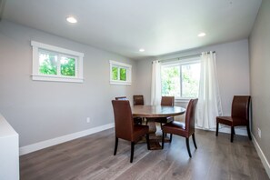 Une salle à manger avec une table ronde, six chaises, deux fenêtres et une vue sur la verdure à l’extérieur.