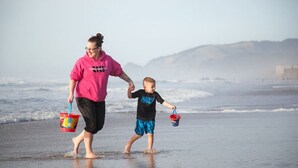 Cena de praia com uma pessoa e uma criança de mãos dadas, ambos carregando baldes.