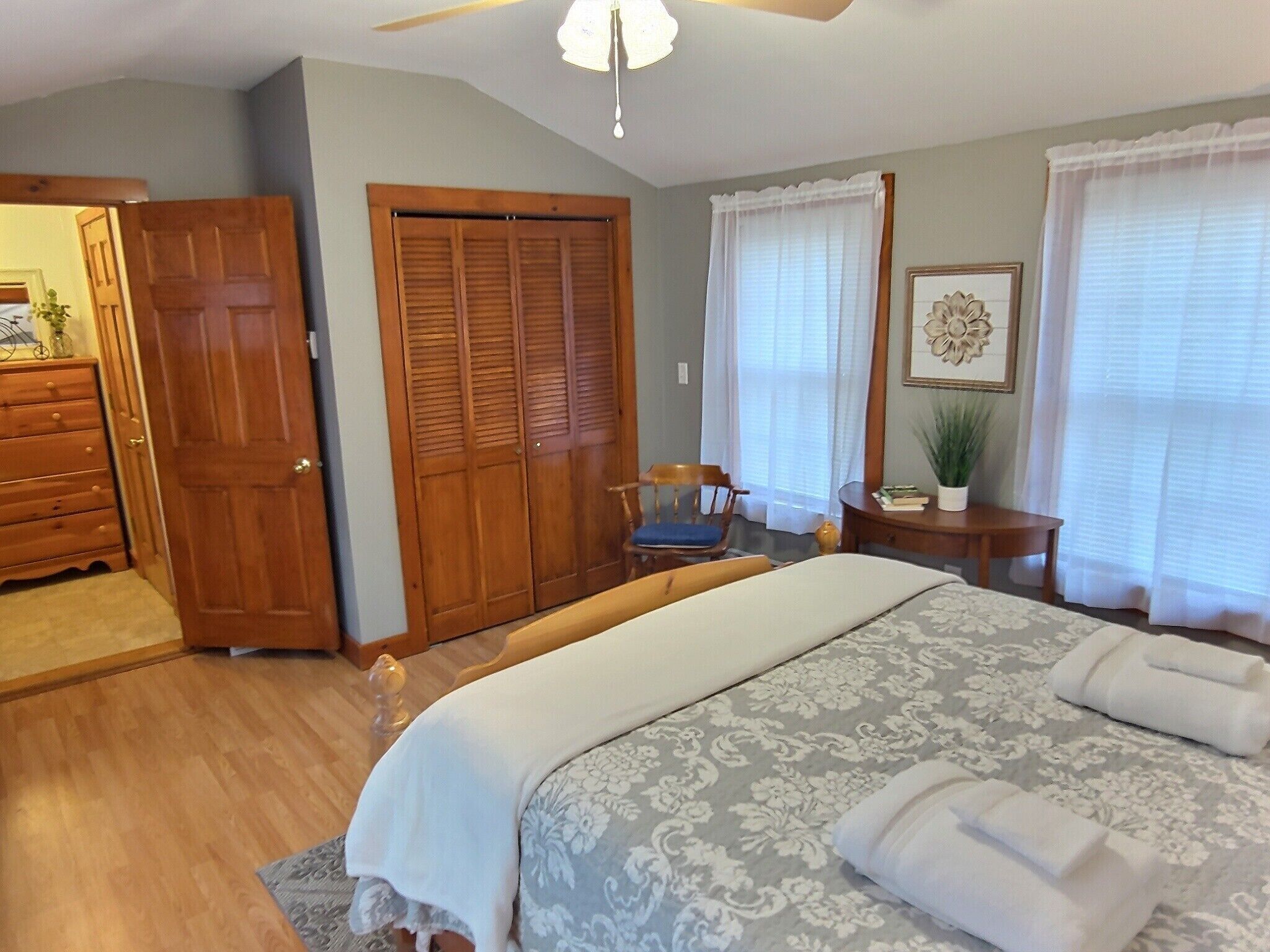 Un dormitorio con una cama, un armario de madera, una silla, una mesita con una planta y una ventana con cortinas.