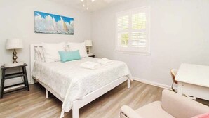Une chambre à coucher avec un lit, un ventilateur de plafond, une fenêtre avec des volets, un tableau et une table de chevet avec une lampe.