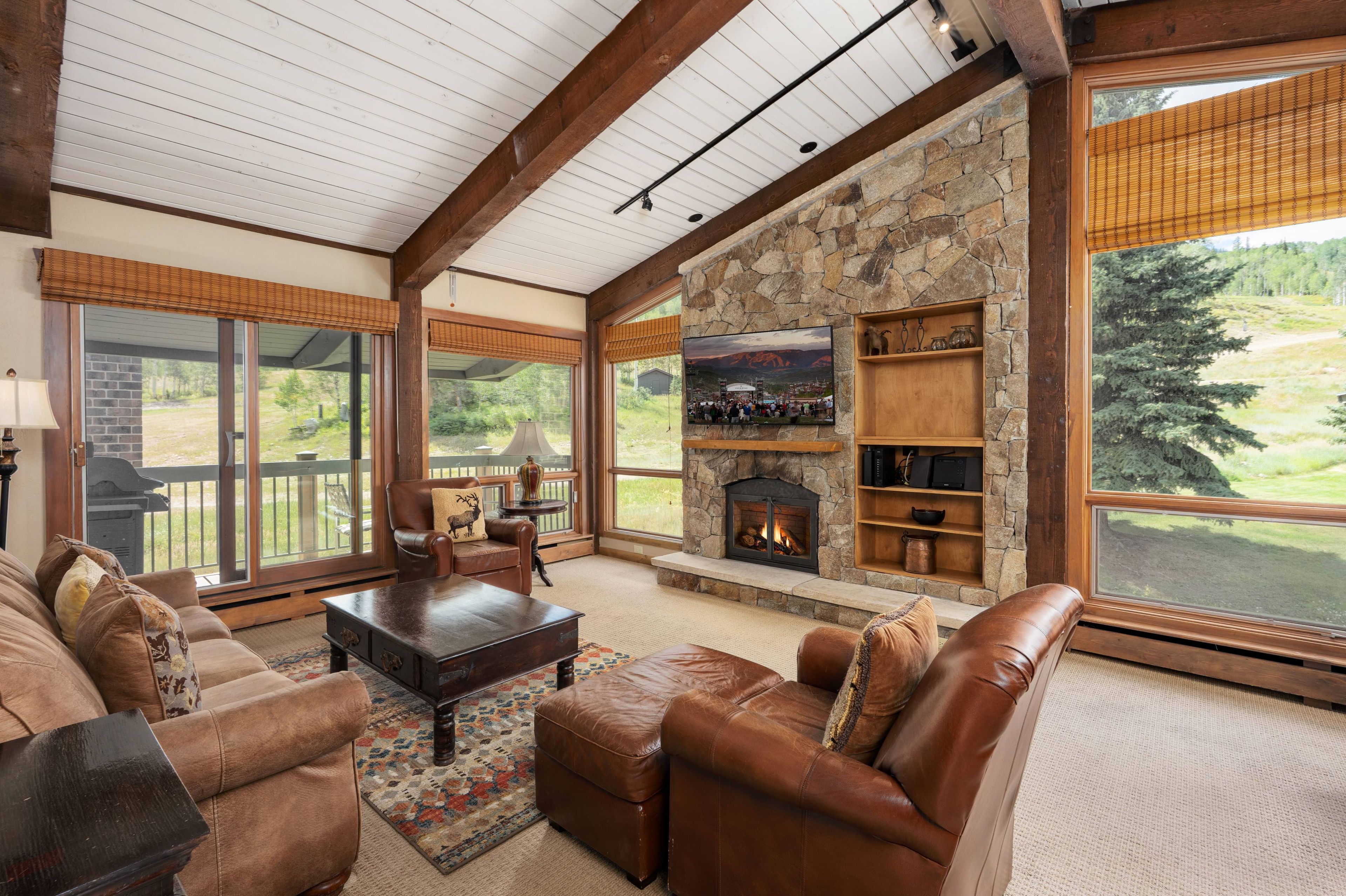 A cozy living room with a stone fireplace, leather furniture, and large windows offering a view of the outdoors.