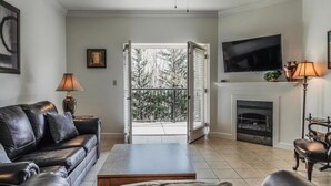 Ein Wohnzimmer mit Sofa, Couchtisch, Kamin, Fernseher und einem Balkon mit Blick auf Bäume.