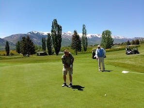 Campo de golf con montañas al fondo, un carrito de golf y una persona preparándose para hacer un putt.