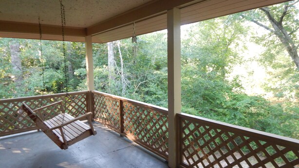 Une véranda couverte avec une balançoire, des balustrades en treillis de bois et une vue sur une zone boisée.