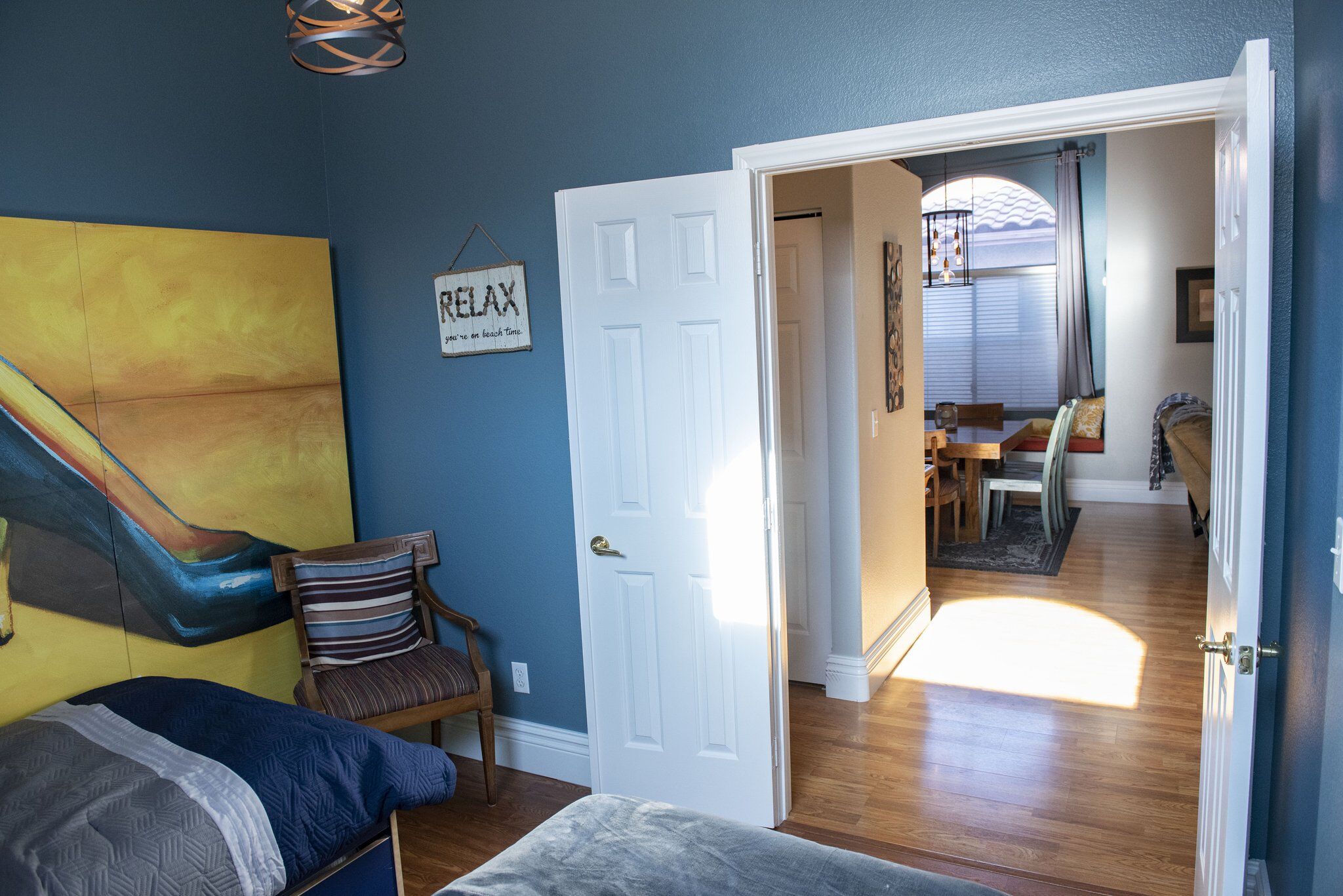 A room with a bed, a chair, a door leading to another room, and a painting on the wall.