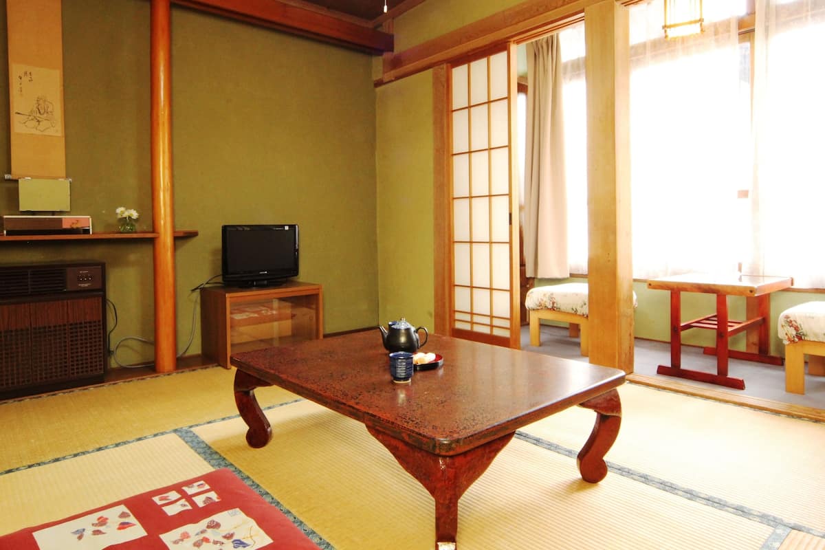 A traditional Japanese room with tatami flooring, a low wooden table, a television on a cabinet, and sliding doors.