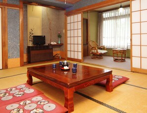 Une pièce de style japonais traditionnel, avec un sol recouvert de tatamis, une table en bois sur laquelle sont posées des tasses et une théière, un téléviseur et une porte coulissante donnant sur une autre pièce.