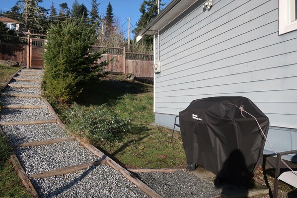 En grussti der fører hen til et hus med grill under tag og et træhegn i baggrunden.