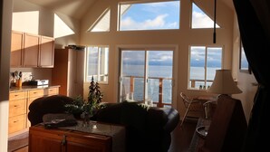 Un'ampia stanza con soffitto alto, grandi finestre vista mare, un'accogliente zona salotto con divano e poltrone, e un mobiletto in legno con elettrodomestici da cucina.