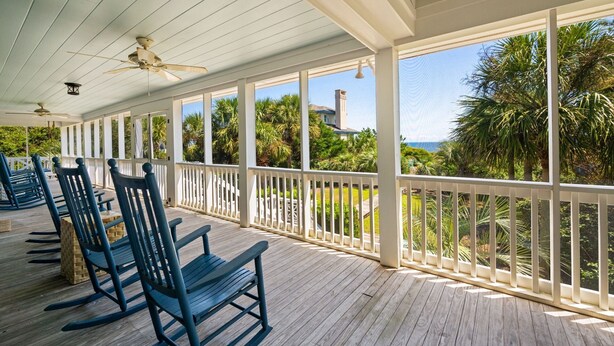 Eine überdachte Veranda mit Holzboden und weißen Geländern, mit Schaukelstühlen und Blick auf ein Haus und Palmen.