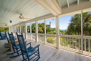 A covered porch with wooden flooring and white railings, featuring rocking chairs and a view of a house and palm trees.