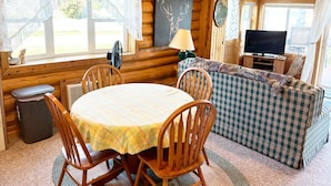 L’intérieur d’un chalet accueillant, avec une table à manger, des chaises, un canapé à carreaux, un téléviseur et une cheminée.