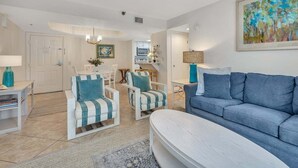 A modern living room with a blue sofa, two striped armchairs, a white coffee table, and a dining area with a white table and chairs. - Oceanfront Gem: Pool, Beach & Prime Location (Saint Simons Island)