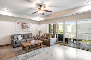 A spacious living room with a ceiling fan, a large window, and a sliding glass door leading to an outdoor patio.