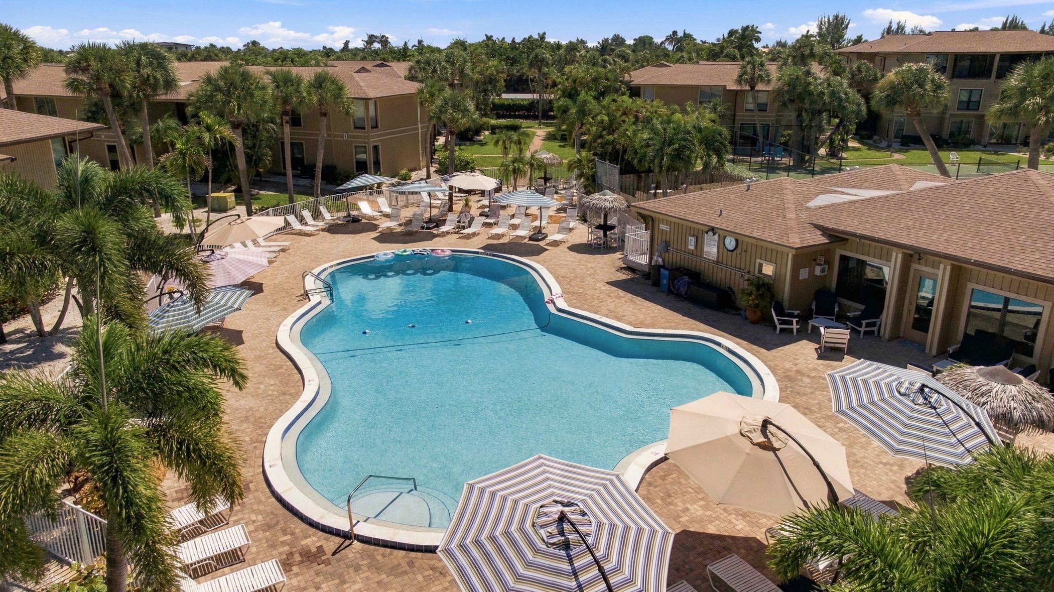 A hotel pool area with a large pool, sun loungers, umbrellas, and palm trees.