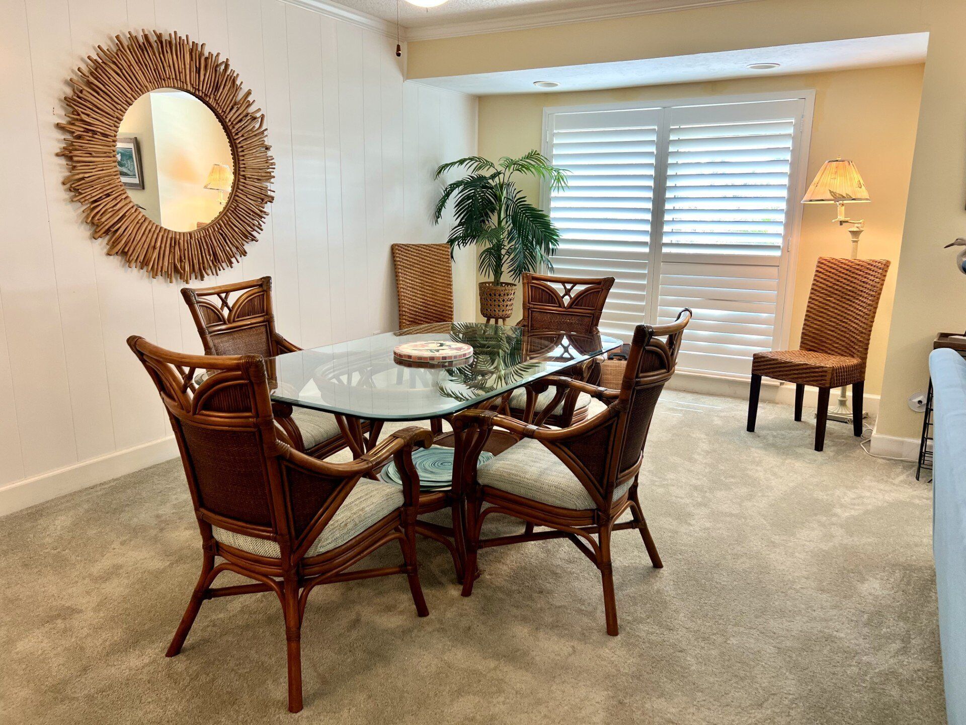 A dining area with a glass table surrounded by wicker chairs, a potted plant, and a wall-mounted mirror.