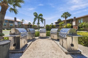 A row of outdoor grills in a courtyard with palm trees and a building in the background.