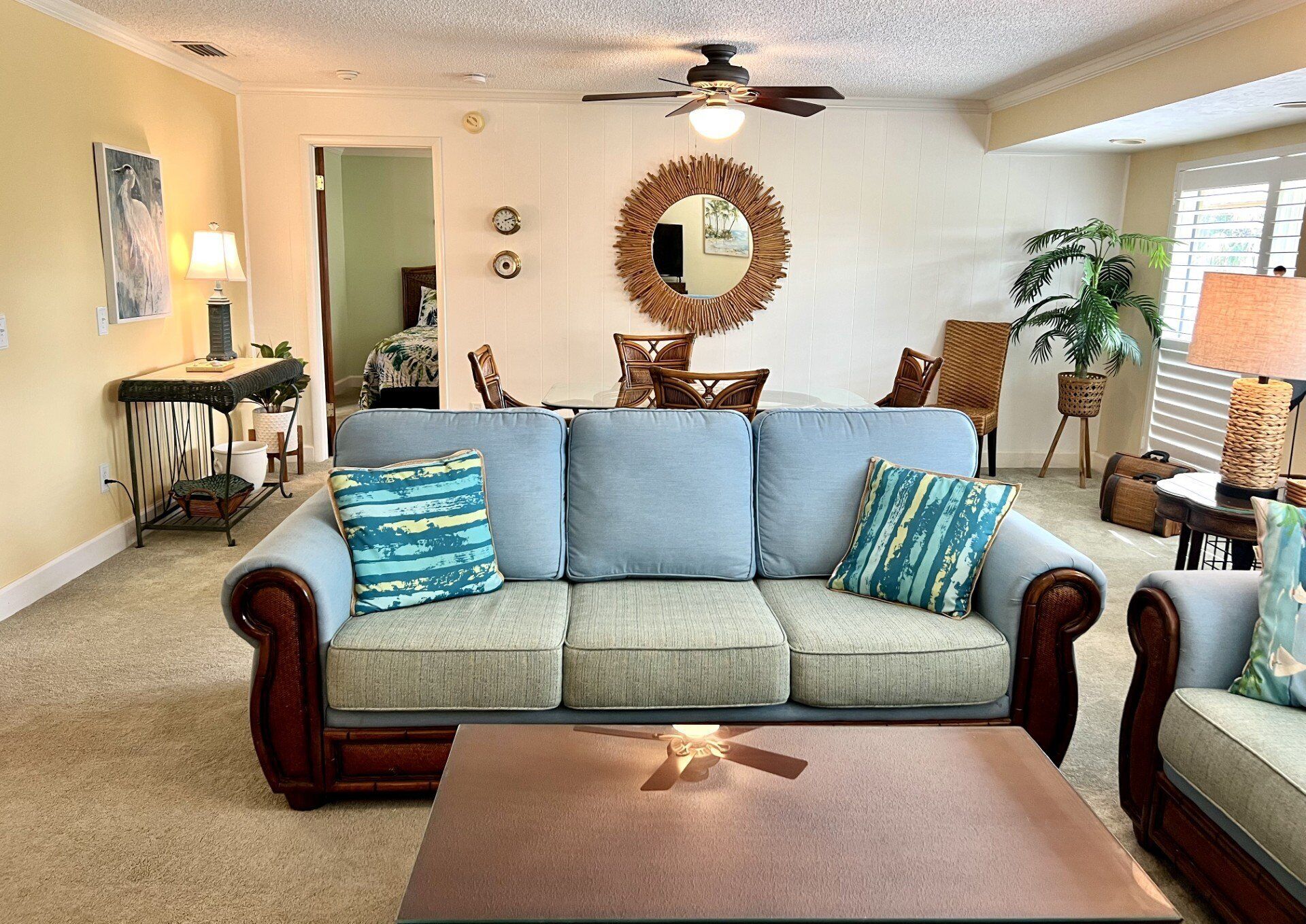 A living room with a sofa, coffee table, and a ceiling fan. There is a dining area with chairs and a table, and a plant in the corner.