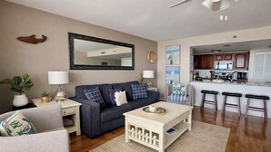 Ein Wohnzimmer mit einem dunkelblauen Sofa, einem weißen Couchtisch und einem grauen Sessel. Ein Küchenbereich mit einer Mikrowelle und einem Spülbecken ist ebenfalls zu sehen.