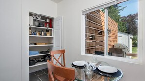 Een compacte keuken met witte kastjes, een eettafel met stoelen en een groot raam met uitzicht op een houten terras en groen.
