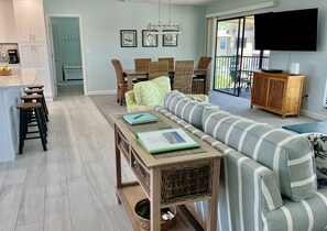 A modern living room with a sofa, a dining table with chairs, a kitchen area with a coffee machine, and a TV mounted on the wall.