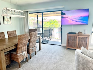 Ein Essbereich mit Korbstühlen und einem Holztisch, einem Fernseher an der Wand und Blick auf einen Strand durch eine Schiebetür.