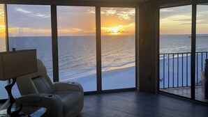 Habitación con un amplio ventanal con vista al mar y al atardecer. Hay un sofá y una mesa con una lámpara.