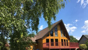 Un'ampia casa in legno con un balcone sporgente, immersa nel verde e sotto un cielo sereno.