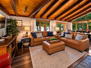 Una sala de estar acogedora con techo de madera, un amplio sofá marrón, una mesa de centro y vistas al exterior a través de amplios ventanales.