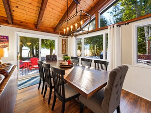 Un comedor con mesa y sillas de madera, una araña y ventanas amplias.