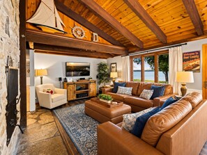 Una sala de estar acogedora con un amplio sofá de cuero marrón, un sillón blanco y un mueble de entretenimiento de madera. Un gran ventanal ofrece vistas al exterior.